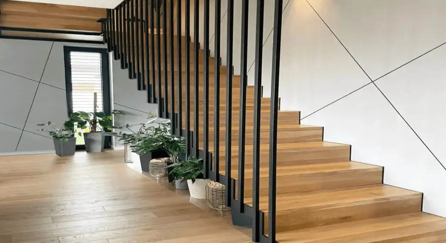 Nowoczesna klatka schodowa w domu z drewnianymi stopniami i czarną, metalową balustradą. Wokół rośliny w doniczkach.