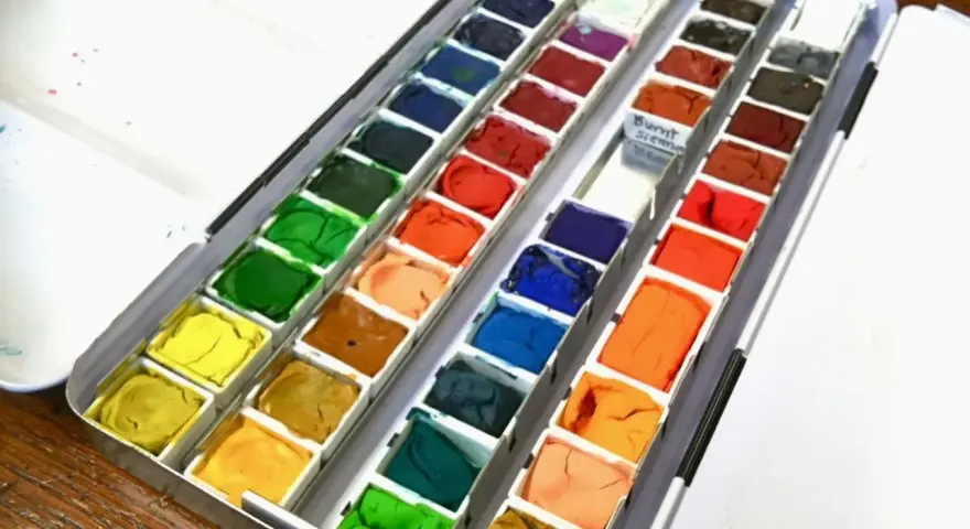 Paleta z różnymi rodzajami farb plastycznych: akwareli w kostkach, od żółci, przez zielenie, po brązy i błękity.