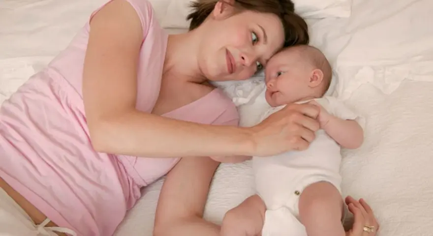Mama patrzy z miłością na swoje 5 tygodniowe dziecko leżące obok niej na łóżku.