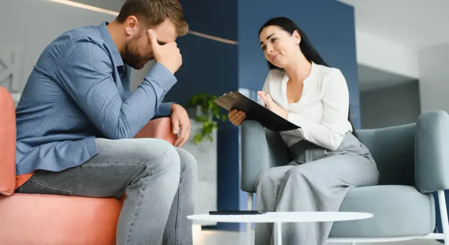 Mężczyzna w stresie podczas sesji, która może być częścią terapii uzależnień online. Terapeutaka notuje.