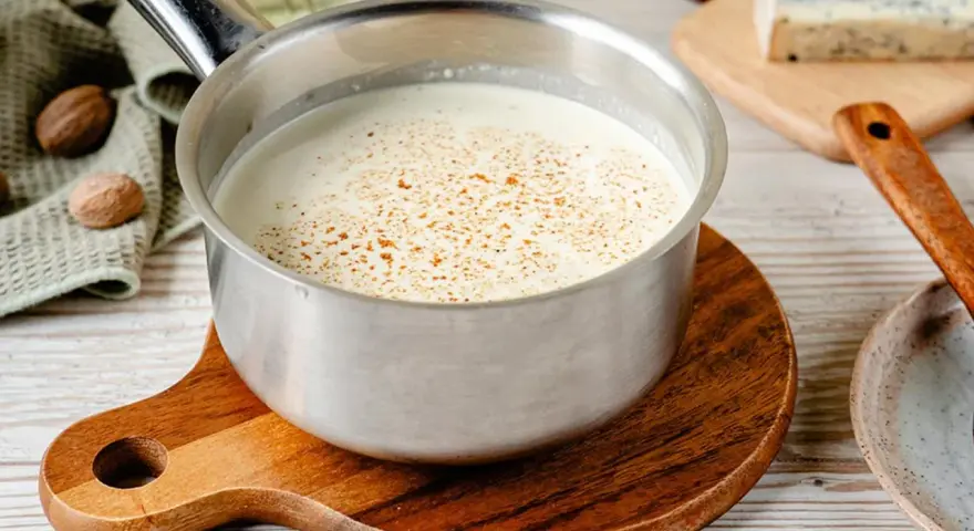 Cremige Gorgonzola-Sauce in einem Topf, bestreut mit Gewürzen. Daneben liegen Muskatnüsse und ein Stück Gorgonzola.