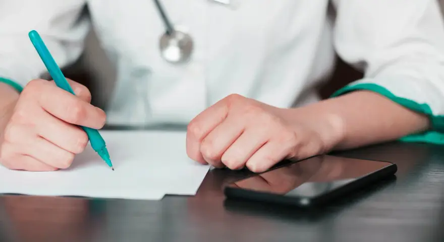 Lekarz pisze zwolnienie, zastanawiając się, na ile lekarz może wystawić L4 jednorazowo. Obok leży telefon.