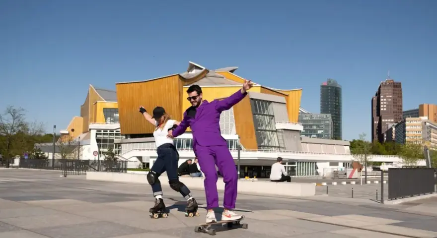 Ein Mann im lila Anzug auf einem Skateboard und eine Frau auf Rollschuhen tanzen vor der Philharmonie. Ein sonniger Tag heute in Berlin.