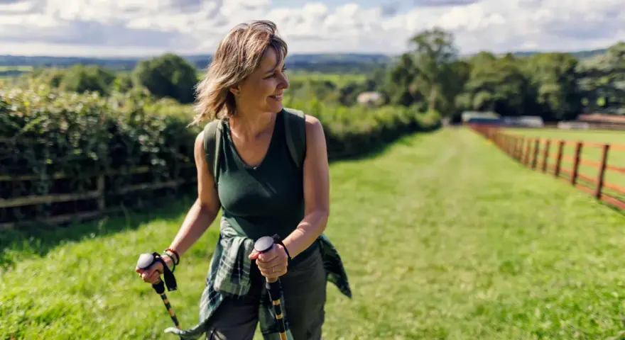 Donna sorridente con bastoncini da nordic walking, che lavora i muscoli coinvolti in un paesaggio verdeggiante.