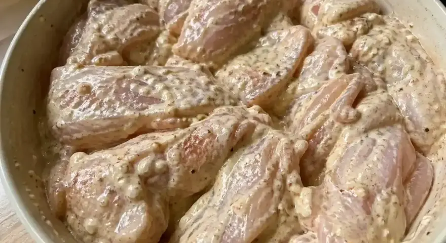 En skål med yoghurtmarinerad kyckling, redo att tillagas. Kycklingbitarna är täckta av en krämig, kryddig marinad.