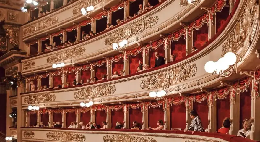Widownia La Scali, pełna widzów w lożach, oczekuje na rozpoczęcie spektaklu. Bogaty repertuar teatru przyciąga miłośników sztuki.