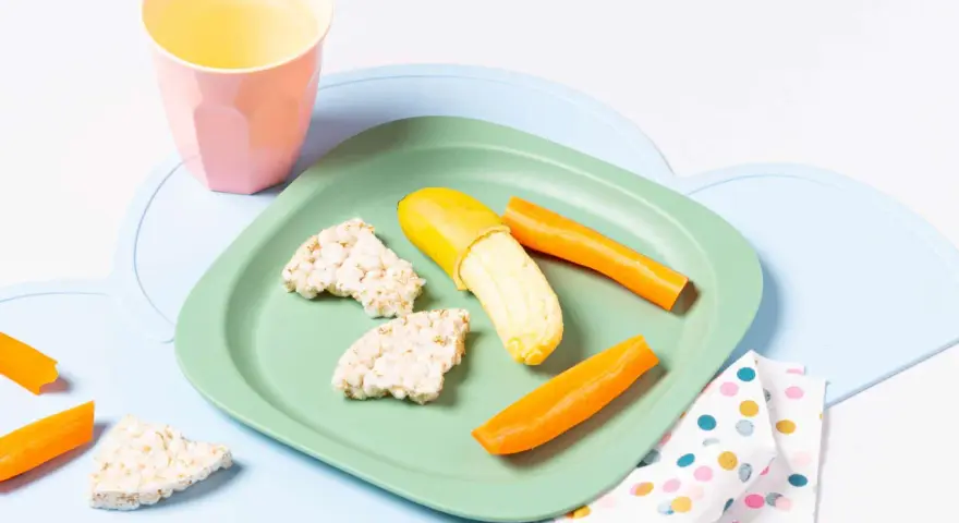 Baby-gerechte Snacks: Reiswaffeln, Karottensticks und eine halbe Banane auf einem grünen Teller.