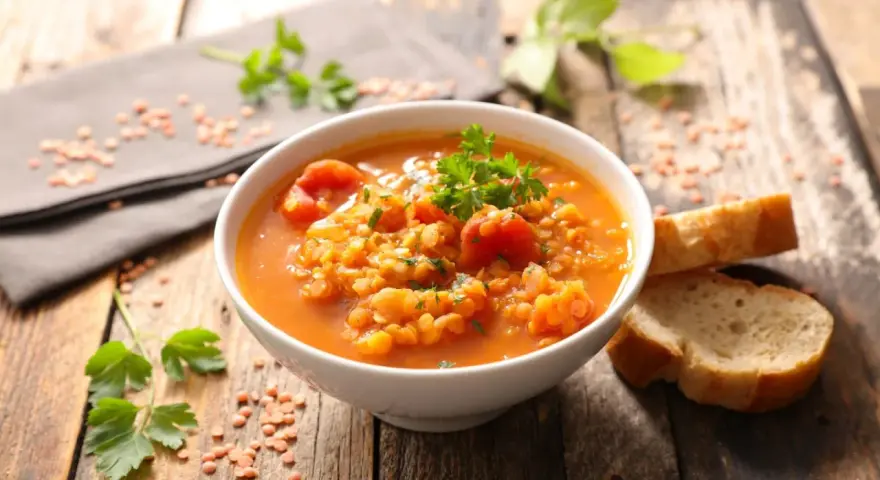 Eine dampfende Schüssel mit herzhafter rote linsensuppe, garniert mit Petersilie, daneben knuspriges Brot auf rustikalem Holztisch.