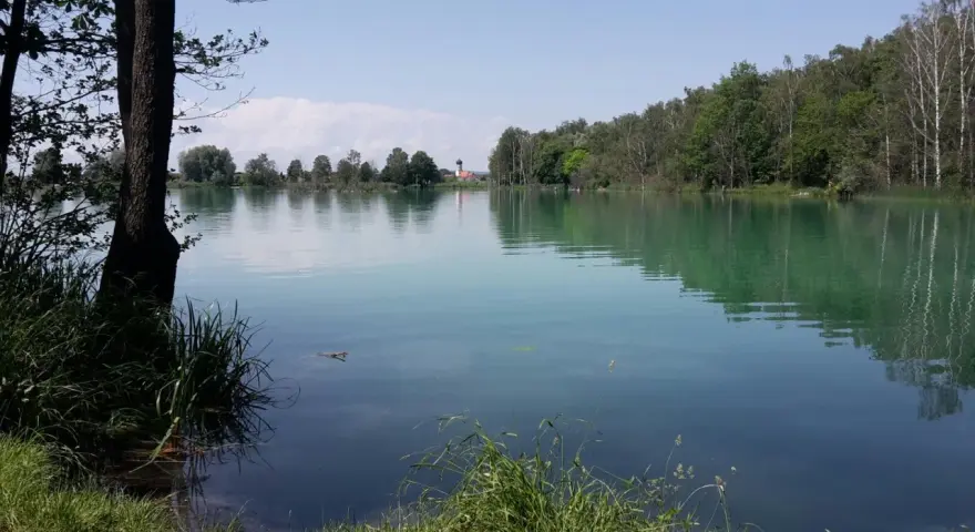 Idyllischer ersinger badesee mit grünen Ufern und einem Kirchturm in der Ferne.