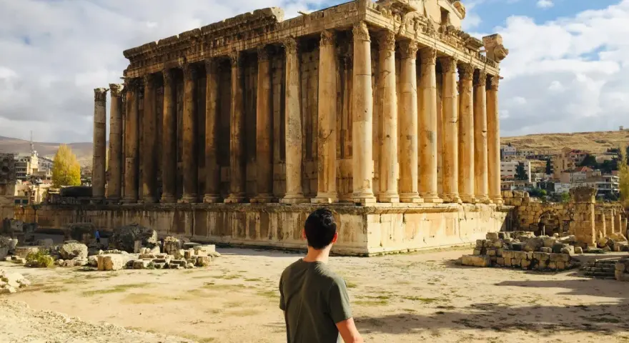 A man sits contemplating the grandeur of Roman ruins in Lebanon, bathed in sunlight under a cloudy sky.