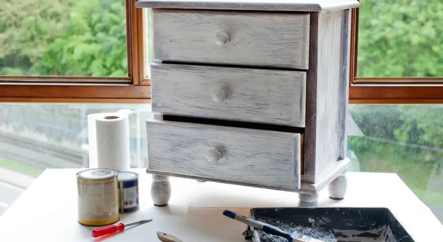 Mueble de tres cajones con acabado desgastado, listo para pintar muebles oscuros. Pinceles, rodillo y latas de pintura a su lado.