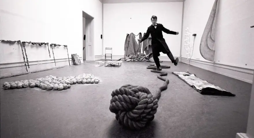 Un homme en costume, peut-être Lawrence Weiner, danse au milieu d'une installation artistique avec des cordes et des objets.