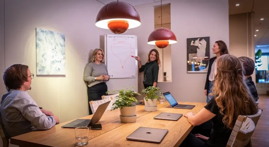 Ett team diskuterar strategier vid ett konferensbord med flera laptops. En kvinna pekar på en trattmodell på en whiteboard. Ups datorn är en del av deras effektiva arbetsflöde.