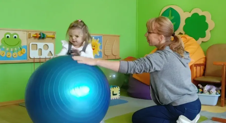 Dziecko w okularach ćwiczy z fizjoterapeutką na dużej, niebieskiej piłce w sali przedszkolnej.