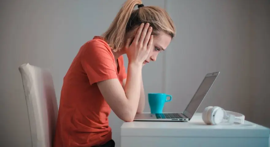 Zmartwiona kobieta przy laptopie, zastanawia się, czy porzucenie pracy wlicza się do emerytury. Na biurku kubek i słuchawki.