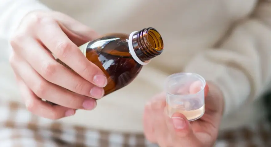 Dłoń przelewa płyn z brązowej butelki do plastikowego kieliszka. To domowy syrop na kaszel, przygotowany z naturalnych składników.
