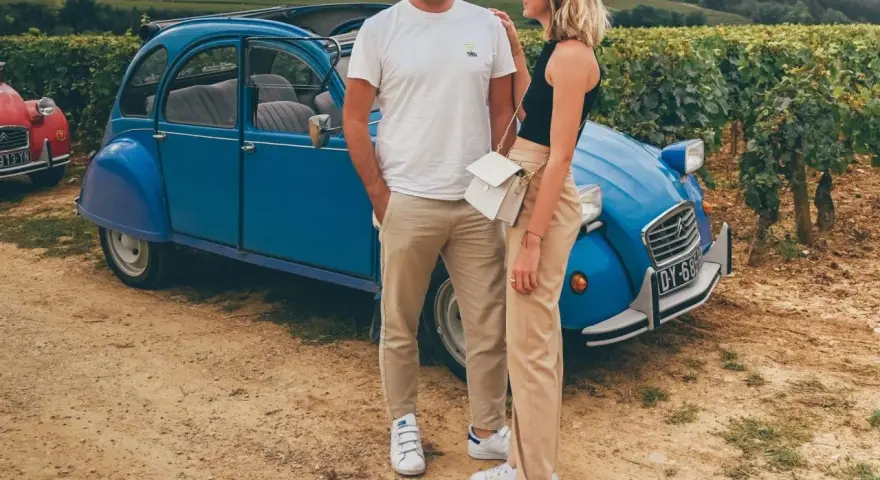 Couple souriant devant une 2CV bleue dans les vignes, une belle idée pour visiter Beaune en 1 jour.