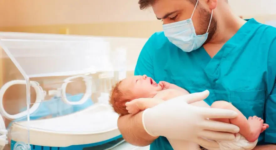 Lekarz w maseczce i rękawiczkach trzyma noworodka. To przykład troski w neonatologii, która zajmuje się opieką nad noworodkami.