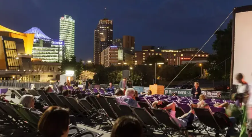 Leute entspannen sich in Liegestühlen beim **Sommerkino am Potsdamer Platz** vor einer leuchtenden Skyline.