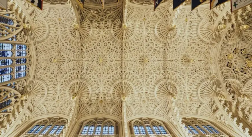 Intricate fan vaulting of a grand hall, reminiscent of the detailed ceilings found in abbey graves, adorned with flags.