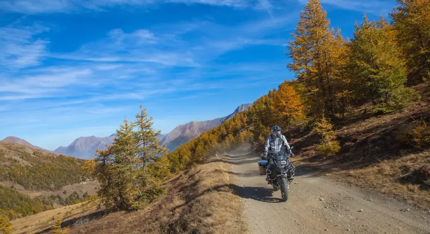 Motociclista su strada sterrata, strada colle delle finestre, con larici dorati e montagne sullo sfondo.