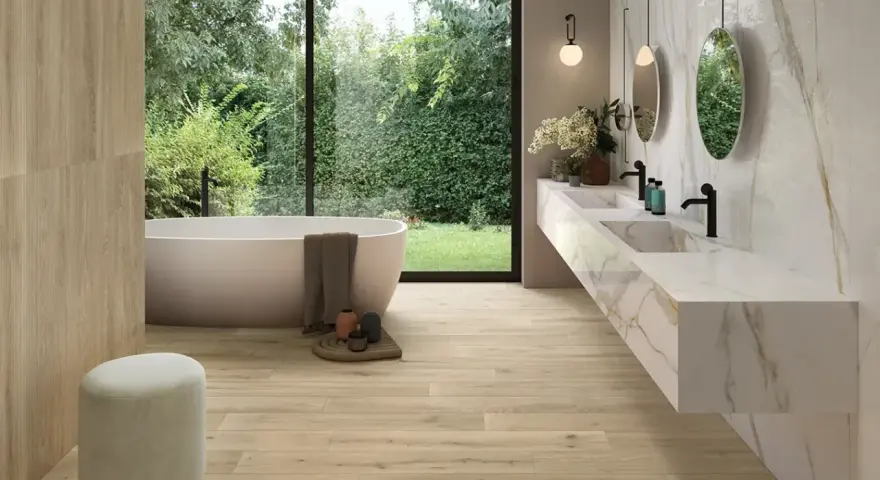 Salle de bain moderne avec baignoire îlot, double vasque en marbre et **pose carrelage imitation parquet** au sol et sur un mur. Vue sur jardin luxuriant.