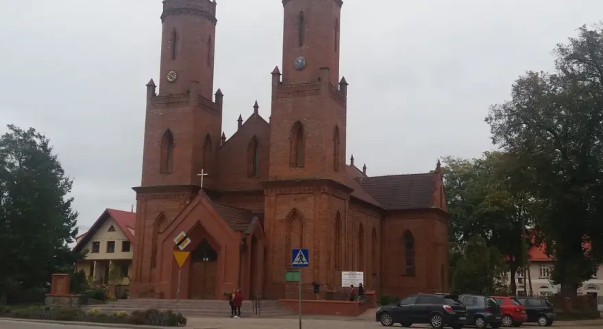 Imponujący kościół w Krokowej z cegły, z dwiema wieżami i zegarami, otoczony zielenią i budynkami.