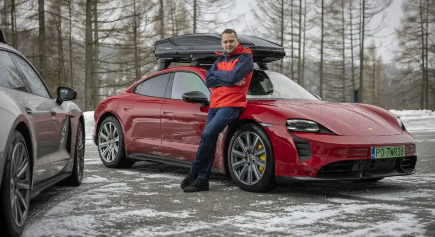Mężczyzna w czerwonej kurtce opiera się o czerwonego Porsche Taycan z boxem dachowym.