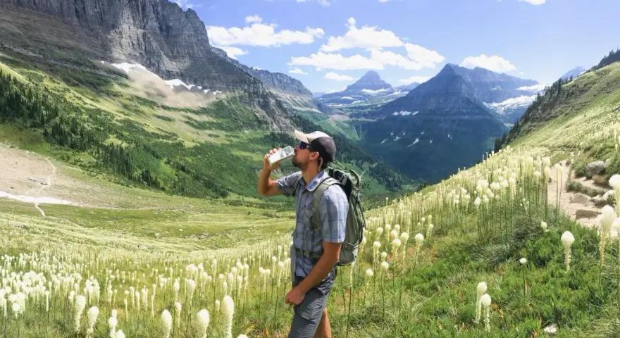 Randonneur buvant de l'eau au milieu d'un champ de fleurs blanches, avec des montagnes majestueuses en arrière-plan. Il espère que cela soulagera sa crampe cuisse.