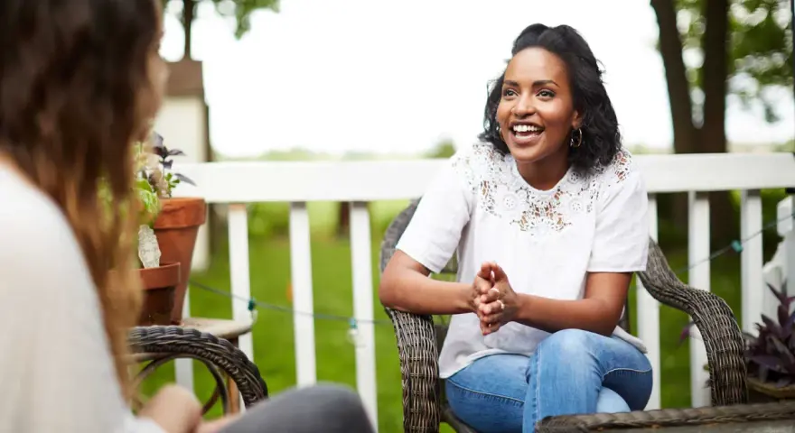 Two women are sitting on a porch, one animatedly sharing the gospel with the other.