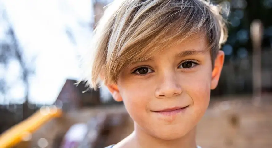 Ein Junge mit blonden Haaren, die nach vorne fallen, lächelt in die Kamera.