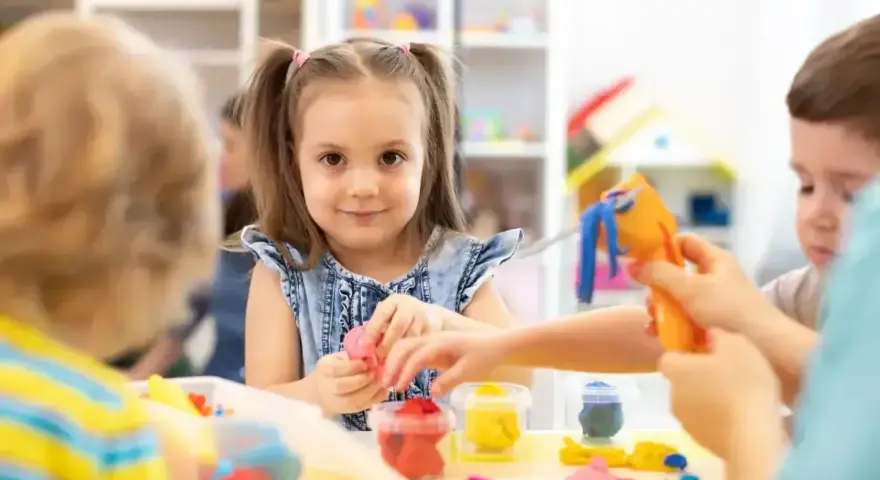 Uśmiechnięta dziewczynka z kitkami bawi się plasteliną, zastanawiając się, w jakim wieku do szkoły pójdzie. Dzieci tworzą kolorowe figurki.