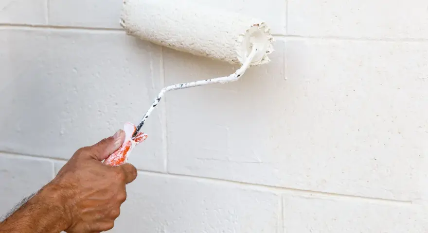 Main tenant, avis peinture carrelage sol : un rouleau blanc applique une nouvelle couche sur un mur en parpaings.