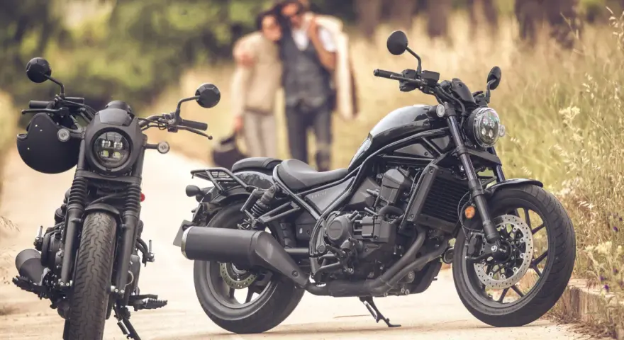 Dos motos negras, una con casco, listas para un viaje. Una pareja abraza en el fondo, disfrutando del paisaje. Una moto sin marchas, perfecta para la aventura.