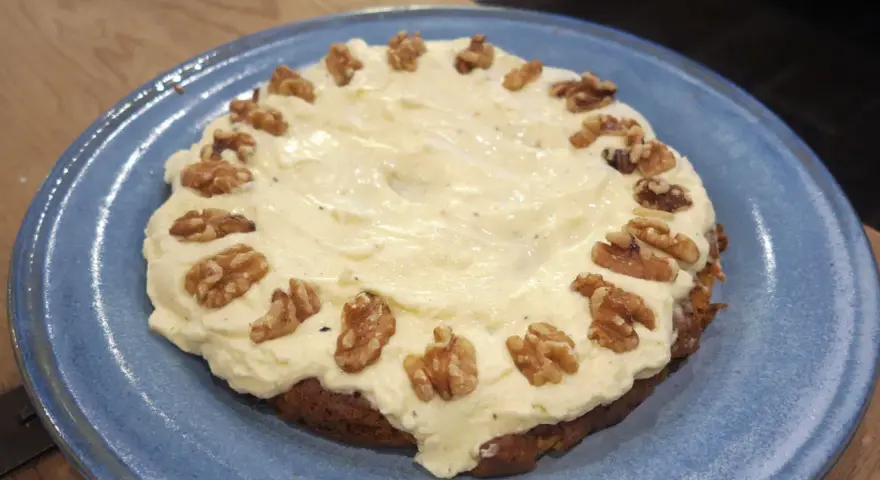 En saftig glutenfri morotskaka med krämig frosting och valnötter på en blå tallrik.