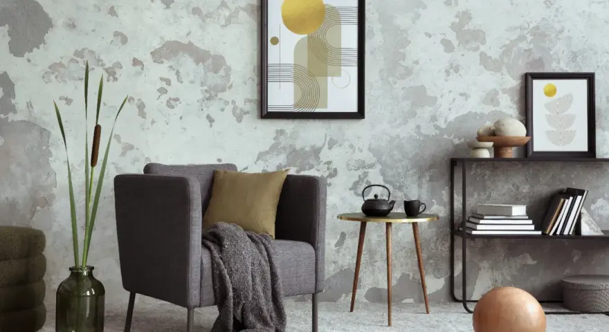 Salón moderno con pared texturizada que combina papel pintado y pintura. Sillón gris, mesa auxiliar dorada, estantería y arte abstracto.