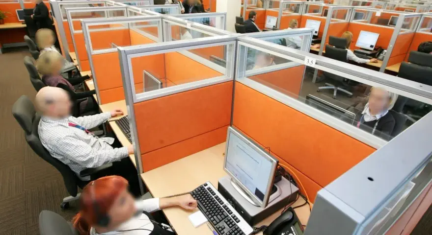 Wielostanowiskowe biuro, gdzie pracownicy obsługują telefony i komputery. W tle widać wiele przegródek, co może sugerować pra group nękanie.