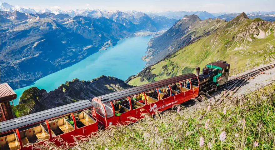 The Brienzer Rothorn train ascends a steep mountain track, offering passengers breathtaking views of a turquoise lake and snow-capped peaks.