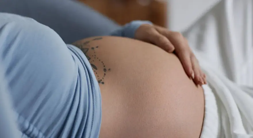 Dłoń spoczywa na **twardym brzuchu w ciąży**, ozdobionym delikatnym tatuażem.