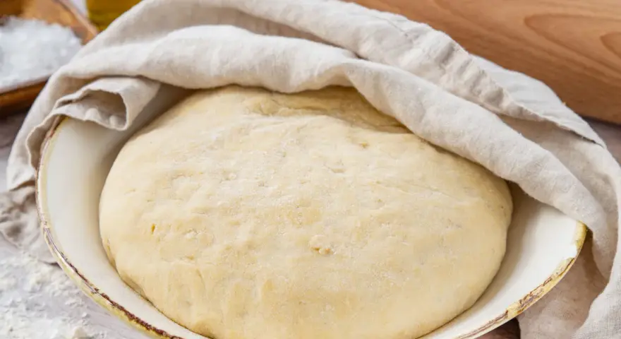 Der weltbeste Pizzateig mit Trockenhefe ruht abgedeckt in einer Schüssel. Daneben liegen Salz, Olivenöl und ein Nudelholz.