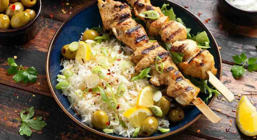 Deliciosos pinchitos de pollo a la parrilla servidos con arroz blanco, aceitunas verdes, rodajas de limón y cebolleta picada.