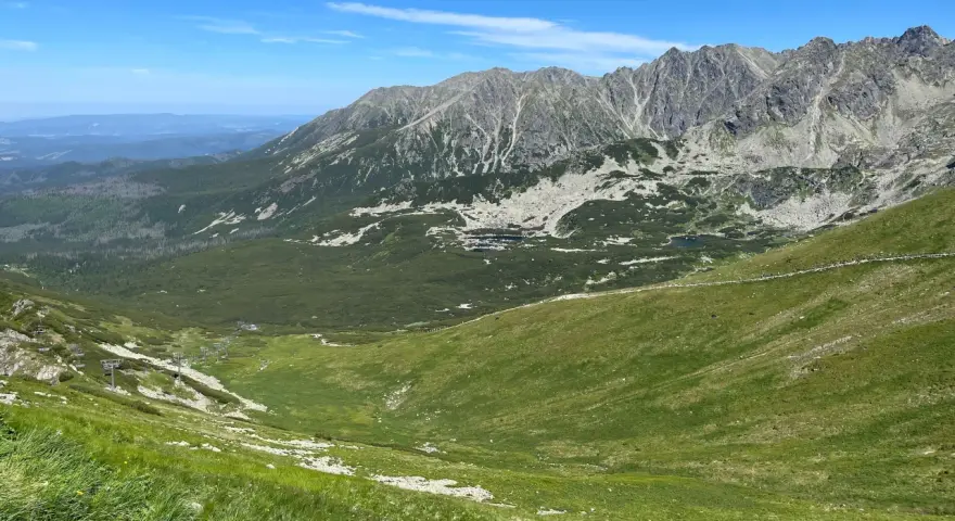 Widok na Tatry z zielonymi zboczami i skalistymi szczytami. W oddali widać budynki. Zastanawiasz się, ile km jest na Kasprowy Wierch?
