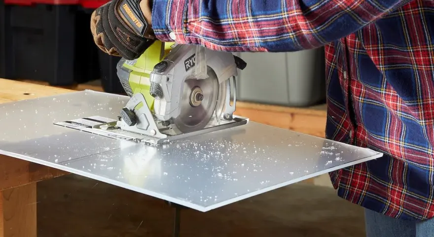 A person in a plaid shirt and gloves uses a circular saw to cut a sheet of plexiglass, creating small plastic shavings. This demonstrates that plexiglass is easy to cut with the right tools.