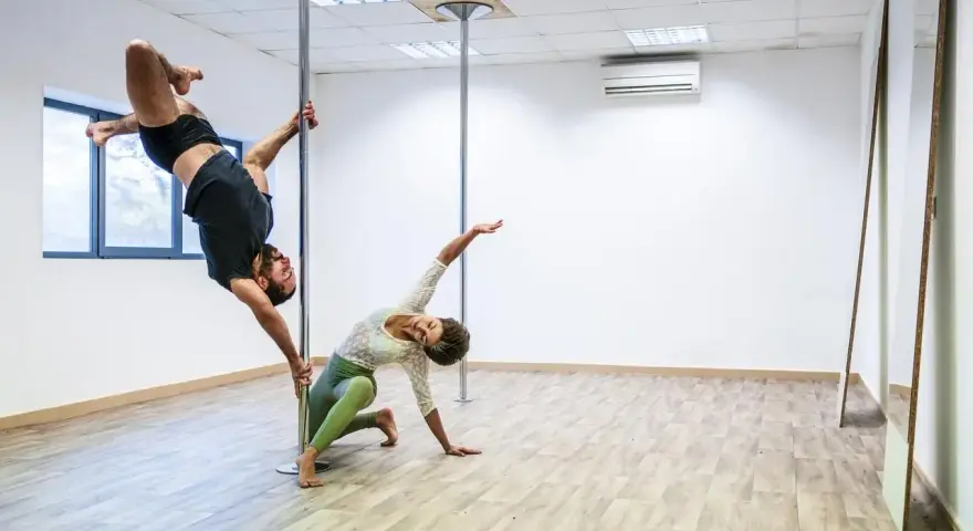 Para ćwiczy pole dance, pokazując efekty odchudzania. On wisi na rurze, ona kuca obok.
