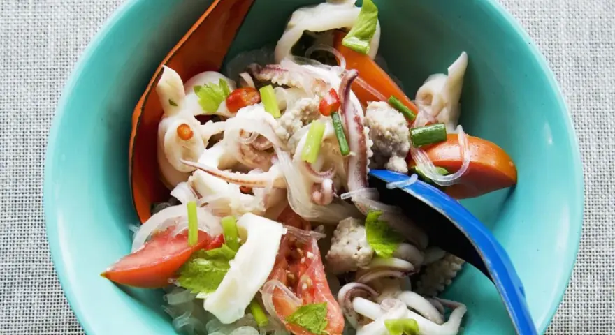 Ensalada tailandesa con fideos de cristal, calamares, tomate y cebolla, servida en un cuenco azul. El plato de sepia en salsa se ve fresco y apetitoso.