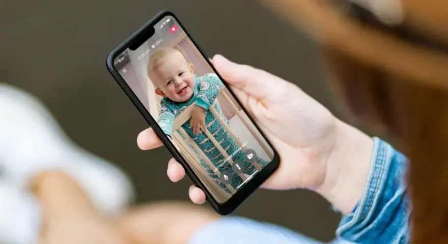 Ein lächelndes Baby im Strampler schaut aus dem Gitterbett. Eine Hand hält das Handy als Babyphone, um das Kind im Blick zu behalten.