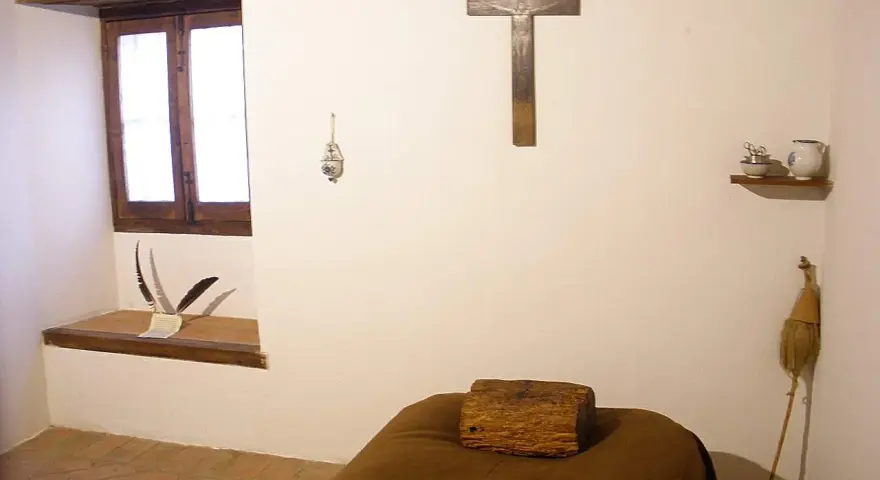 A monk's simple cell, showing where monks sleep: a narrow bed with a wooden block for a pillow, a crucifix, and a window.