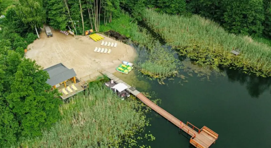 Plaża i pomost nad jeziorem, idealne miejsce na wypoczynek na Pojezierzu Brodnickim. Widać łódki, leżaki i budynek zadaszony.