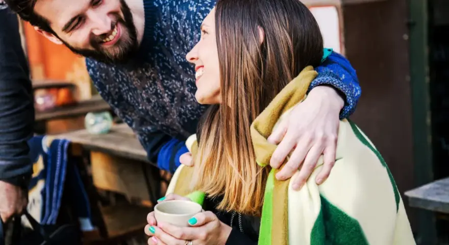 Pareja sonriente compartiendo una manta y una taza de té. Él le abraza por detrás. Ella le dice "muchas gracias en francés".