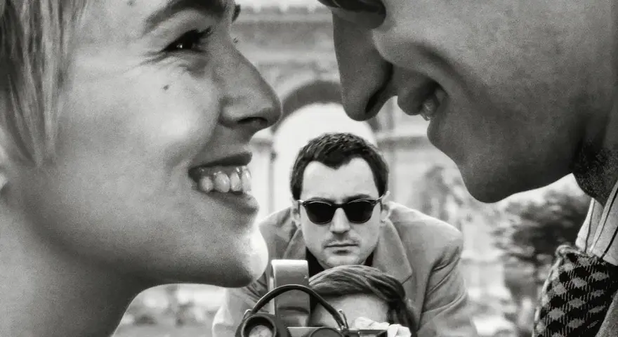 Primo piano di una coppia che ride, con un uomo che filma sullo sfondo, evocando l'estetica dei registi della nouvelle vague.